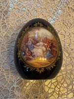 Porcelain egg showing classical painted scene of people in historic clothing with gold floral border on black background, lying flat on woven tablecloth.
