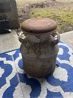Front view of the vintage milk jug placed on a blue and white patterned mat. Shows rust on lid and body.