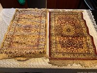 Two small Persian style praying mats laid side by side on a white/cream textured cloth surface, show intricate floral patterns with fringed tassels. One darker red and gold, one yellow and gold. Both appear worn and soiled.