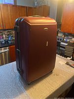 Reddish-brown Samsonite hard shell suitcase standing on a kitchen counter showing front and one side with handle and wheels visible.
