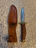 Photo showing the Ruko Cutter knife with its brown leather sheath beside it, placed on a textured beige surface.