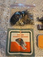 Shows Singer Automatic Zigzagger attachment in plastic bag along with original instruction card and additional small yellow container.