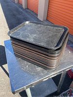 Stack of black and brown rectangular plastic trays with raised edges and indentations on top, placed on metallic table.