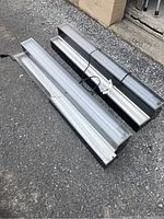 Two LED light shelves placed on a gravel surface. One shelf visible with black casing and silver light panel; the other with silver casing and white light panel damaged at one corner. Both have power cords.