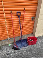 Photo showing five cleaning and yard tools including wood stick rake, two red brooms, blue snow shovel, red bucket with cleaning accessories.