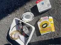 Top-down photo showing a white container filled with assorted plumbing parts including flange rings and seals, along with some packaged items on the ground nearby.