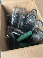 Open cardboard box filled with multiple clear pint glasses featuring Angry Orchard branding, along with green plastic holders for glasses.