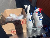 Cardboard box containing loose empty spray bottle containers next to a gray plastic holder containing six spray bottles with colored triggers (blue, red, white).