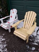 Pair of wooden Muskoka chairs outside on pavement with snow patches, yellow chair on right and decorated white chair on left