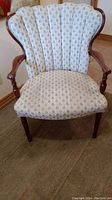 Front view of antique wooden armchair with cream fabric upholstery decorated with small floral patterns, showing the curved wooden arms and scalloped backrest.
