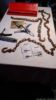 Photo showing orange case, long rusty chains with large hooks, 3-jaw puller, grease squirter, and spark plugs arranged on table.