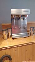 Front view of stainless steel mixer with three spindles and three cups on wooden surface