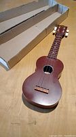 Full ukulele with original cardboard box on wooden surface.