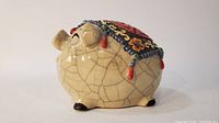 Side view of cream-colored porcelain turtle bank with crackle glaze, detailed molded facial and foot features, colorful painted shell with hanging bead accents.