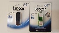 Front view of two Lexar 64GB JumpDrive USB flash drives in unopened packaging, one USB 2.0 model S70 in black, one USB 3.0 model S75 in green and white.