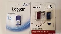 Front view of Lexar 64GB JumpDrive V30 USB 2.0 flash drive package and PNY 16GB USB Flash Drive 2 pack package showing both unopened in original packaging.