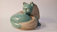 View of the cat figurine showing its greenish teal glazed face and body with textured beige tail wrapped around.