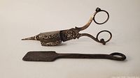 Two items shown: a silver plated candle wick cutter with ornate floral detailing and footed base, and a flat iron tool with a looped handle.