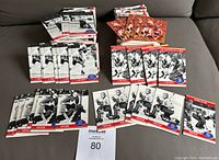 Photo showing multiple stacks of hockey cards from the 1972 Canada Russia Series, black and white photos with red and blue accents, placed on a gray couch surface with a MaxVida card and auction card number 80 visible.