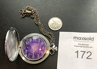 Photo of vintage pocket watch with purple face, open cover, chain and a quarter coin for size comparison.
