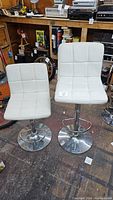 Two white bar stools on a wooden floor with visible pads and chrome metal bases. Background includes various tools and items on shelves.