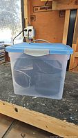 Clear plastic storage container with blue lid, holding various camping items inside, placed on workbench with wood background.