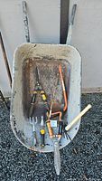 Photo showing all garden tools arranged inside metal wheelbarrow including buck saw, pruning shears, shears, hand fork, cultivator, and small rake.