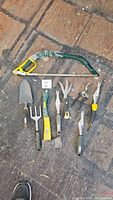 Top view of eight gardening hand tools arranged on floor showing pruning saw, trowel, cultivator, hatchet, and other tools.