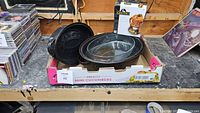 Two black roasting pans with lids and a clear oval glass roasting dish in a cardboard box on a wooden table, with a boxed metal chicken roasting rack beside them.