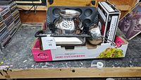 Box holding assorted kitchenware including metal bowls, metal strainer, grill press, and a mini muffin mold.