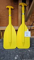 Two yellow plastic paddles with T-shaped handles and adjustable shafts shown side-by-side on a wooden background