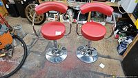Two red bar stools with round padded seats and curved backrests on chrome pedestal bases shown together.