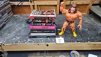 Lot photos showing a stack of 8 wrestling books including biographies and story collections, next to a Warrior action figure with face paint and colorful attire.