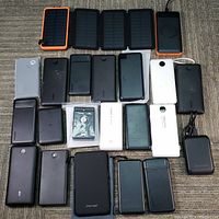 Overview of 20 power banks with black and white finishes in different sizes including some with wireless charging surface and one paw print logo. Foldable solar panels in orange and black frames placed at top.