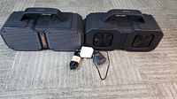 Two large black Bugani M83 portable Bluetooth speakers side by side, with two chargers in front.
