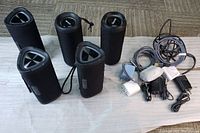 Five black portable speakers with fabric covers, placed on a cloth with several chargers and wires beside them.
