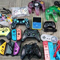 Top down photo showing multiple controllers including Nintendo 64 translucent purple and green, colorful and blue accented PlayStation-style controllers, several Nintendo Switch Joy-Con controllers (red, blue, yellow, purple), Xbox wired controllers, and handheld LCD game device in blue and yellow.