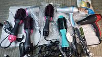 Four Revlon round hair dryers styled brushes with black bristles and colorful handles, two hair dryers, a small hair straightener, and a Pedi Egg foot care product arranged on a fabric surface.
