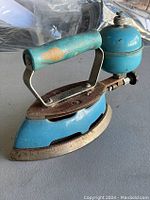 Side angle view of blue Coleman antique iron showing handle, fuel tank, and soleplate with rust and wear.