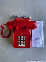Red Cortelco corded desk phone with keypad and handset with coiled cord. User manual for 2500/2554 telephones next to phone.