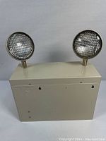 Front view of emergency exit light unit showing two round adjustable light heads on rectangular beige metal housing.