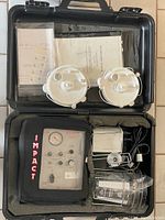 Open carrying case showing Impact 326/326M pump unit, two white diaphragm pump components, tubing, a transparent container, metal clamps, and manuals stored in lid compartment.