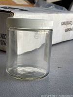 Close-up photo of a single clear glass container with white plastic lid showing its cylindrical shape and flat base.