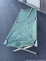 Folding camping cot laid out on dark asphalt surface with metal frame and khaki green fabric surface visible, showing creases from prior use. One metal cross leg visible with black rubber feet caps.