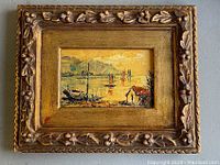 Front view of the small framed vintage painting showing a harbor scene with boats and a small house, ornate carved gold-tone frame.