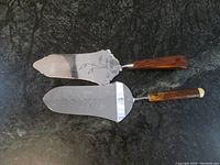 Top-down view of both vintage cake servers showing engraved blades and amber Bakelite handles on dark surface.