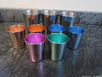 All nine metal drinking cups arranged in two rows, three stainless steel WMF Cromargan cups in back and six colored cups in front.