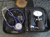 Full view of the open black zippered case containing a purple Littmann stethoscope on the left and a blue SK Spacelab blood pressure cuff with attached inflation bulb and gauge on the right.