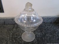 Full view of the clear glass candy dish with a lid featuring a frosted lion figurine on top, standing on a scalloped edge rim dish on a frosted stem and foot.
