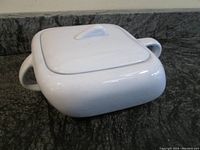 Side angle view of white ceramic casserole dish with lid and side handles, showing the elongated parallelogram shape.